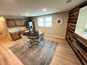A kitchen with wooden cabinets, a refrigerator, a dining table with chairs, and a clock on the wall.