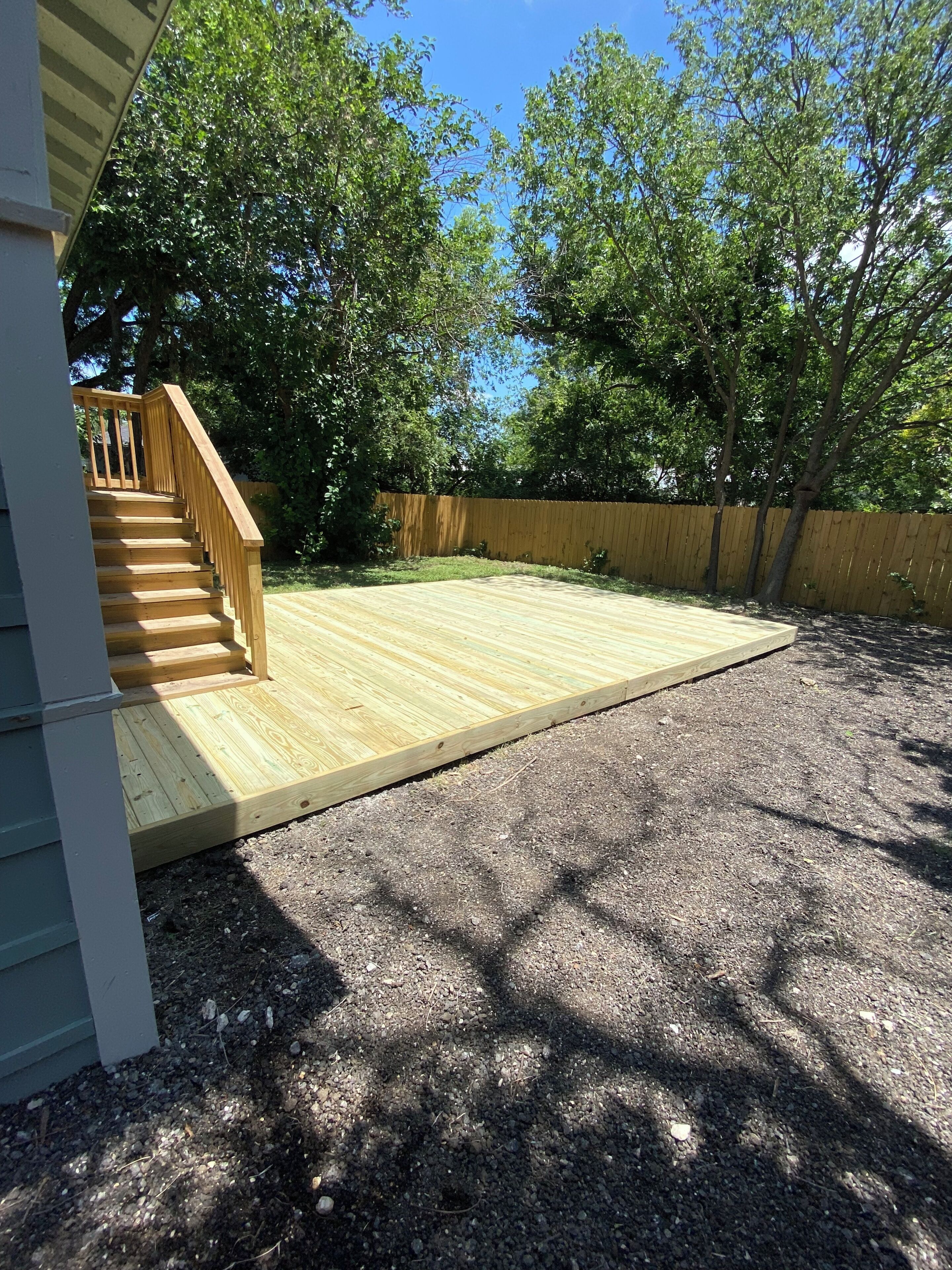 Una terraza de madera con una escalera que lleva a una casa, rodeada de árboles y una cerca de madera.