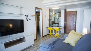 Moderni hotellihuone, jossa on taulutelevisio, tyynyillä varustettu sänky, pieni oleskelualue, jossa on keltaisia tuoleja, ja taustalla näkyvä pieni keittiö.