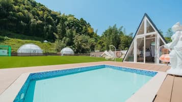 Piscina com borda azul translúcida, rodeada por um deck de madeira. Ao fundo, destacam-se duas estruturas em formato de cúpula e um edifício triangular com fachada de vidro.