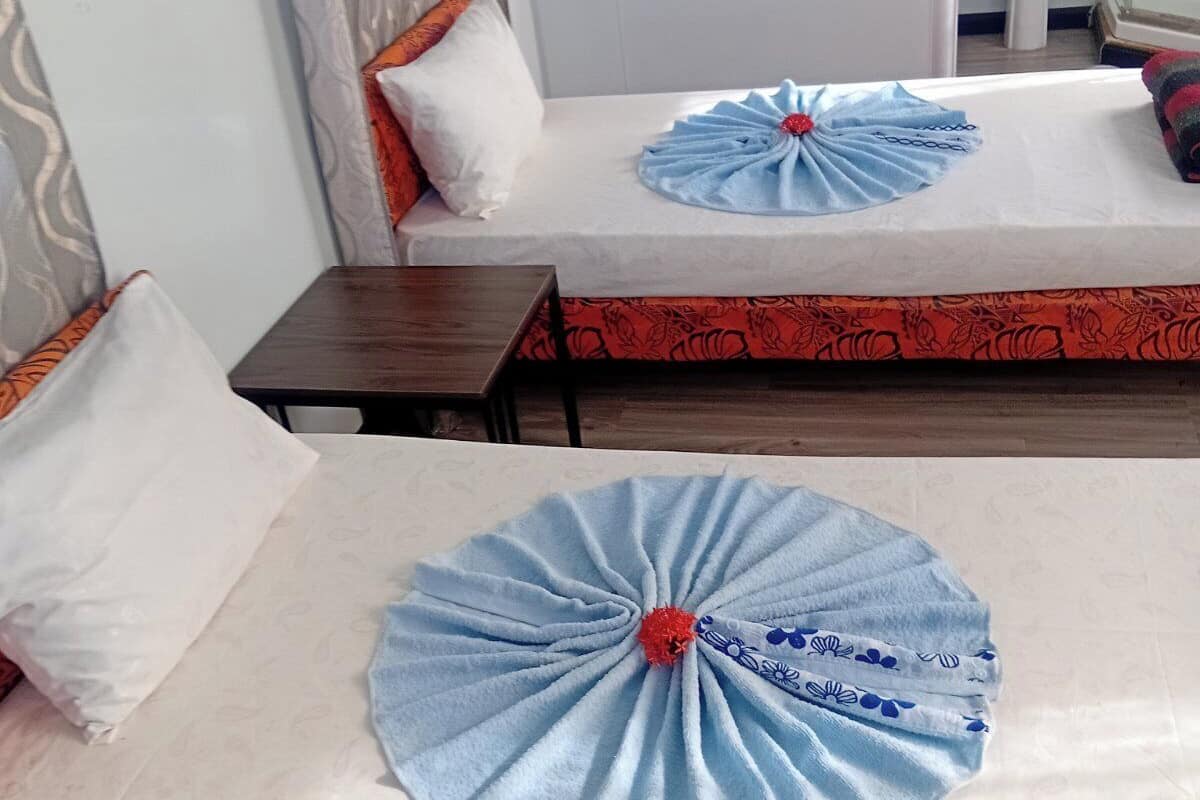 A hotel room with two beds, each with a neatly folded blue towel and a red flower centerpiece. There is a small wooden table and a white sink in the corner.