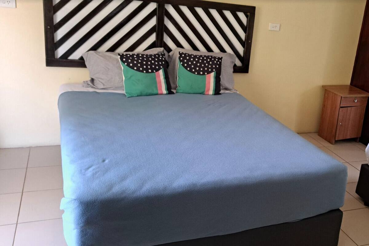 A neatly made bed with a blue bedspread, two pillows with polka dots and stripes, and a wooden nightstand with a drawer.