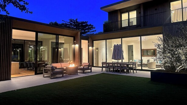 Una casa moderna de dos pisos con un patio que incluye una fogata y mobiliario exterior.