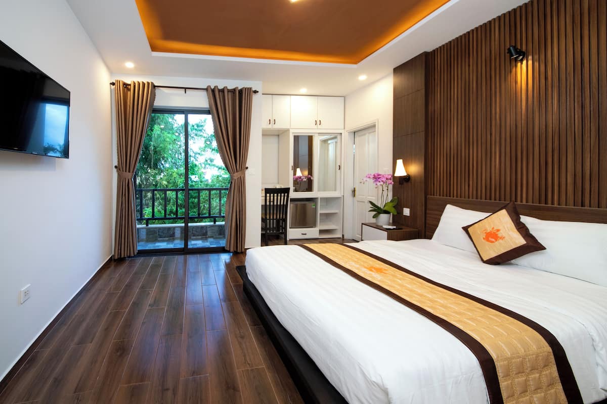 A hotel room with a large bed, a TV mounted on the wall, a balcony with curtains, and a wooden ceiling.