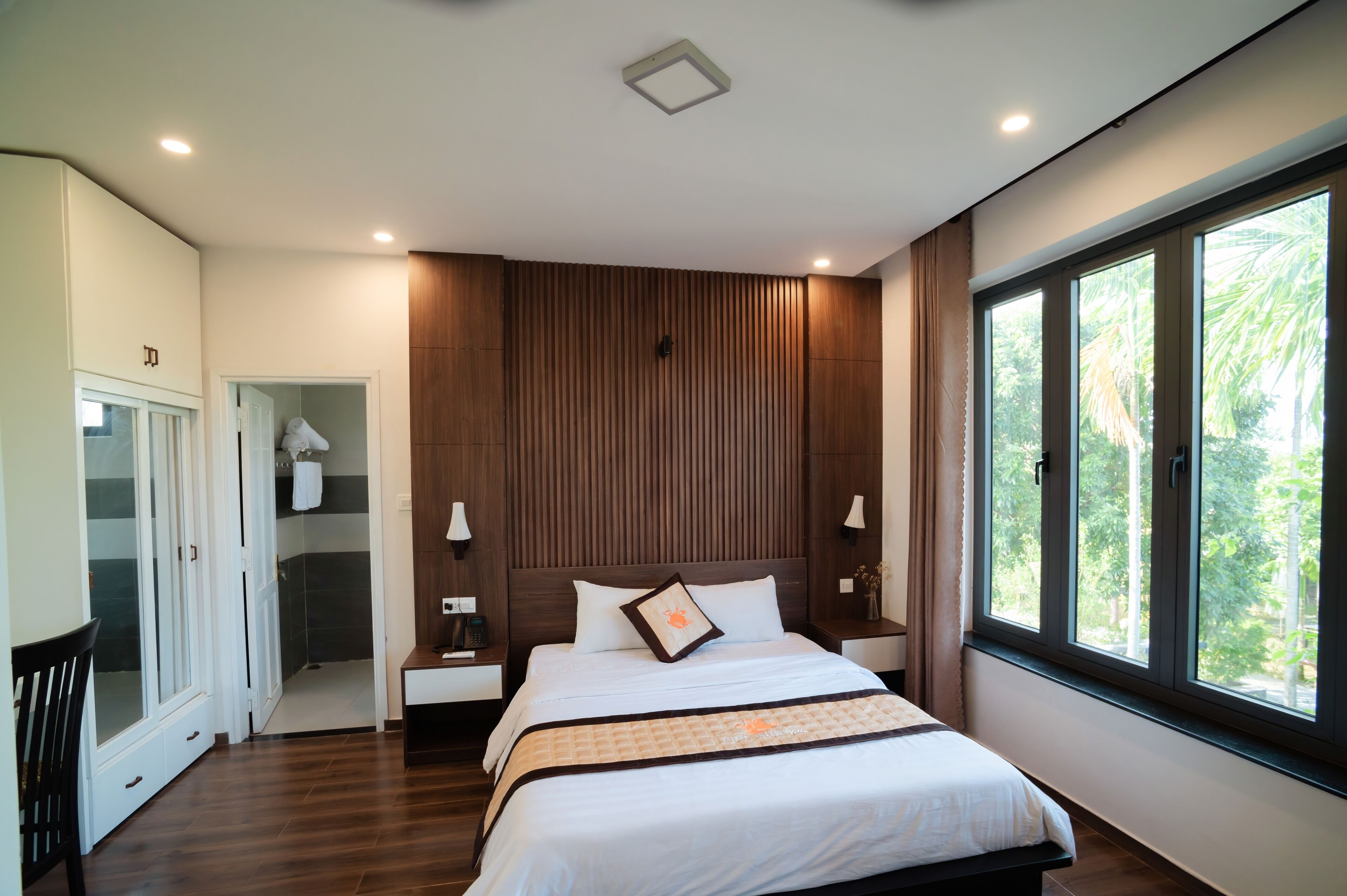 A modern bedroom with a large bed, wooden panel wall, and a window with a view of greenery.