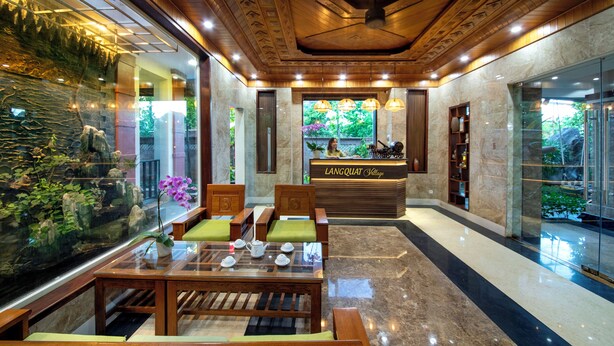 Un área de recepción con mostrador de madera, zona de estar con sillas y una mesa, un acuario y vistas al exterior con vegetación.