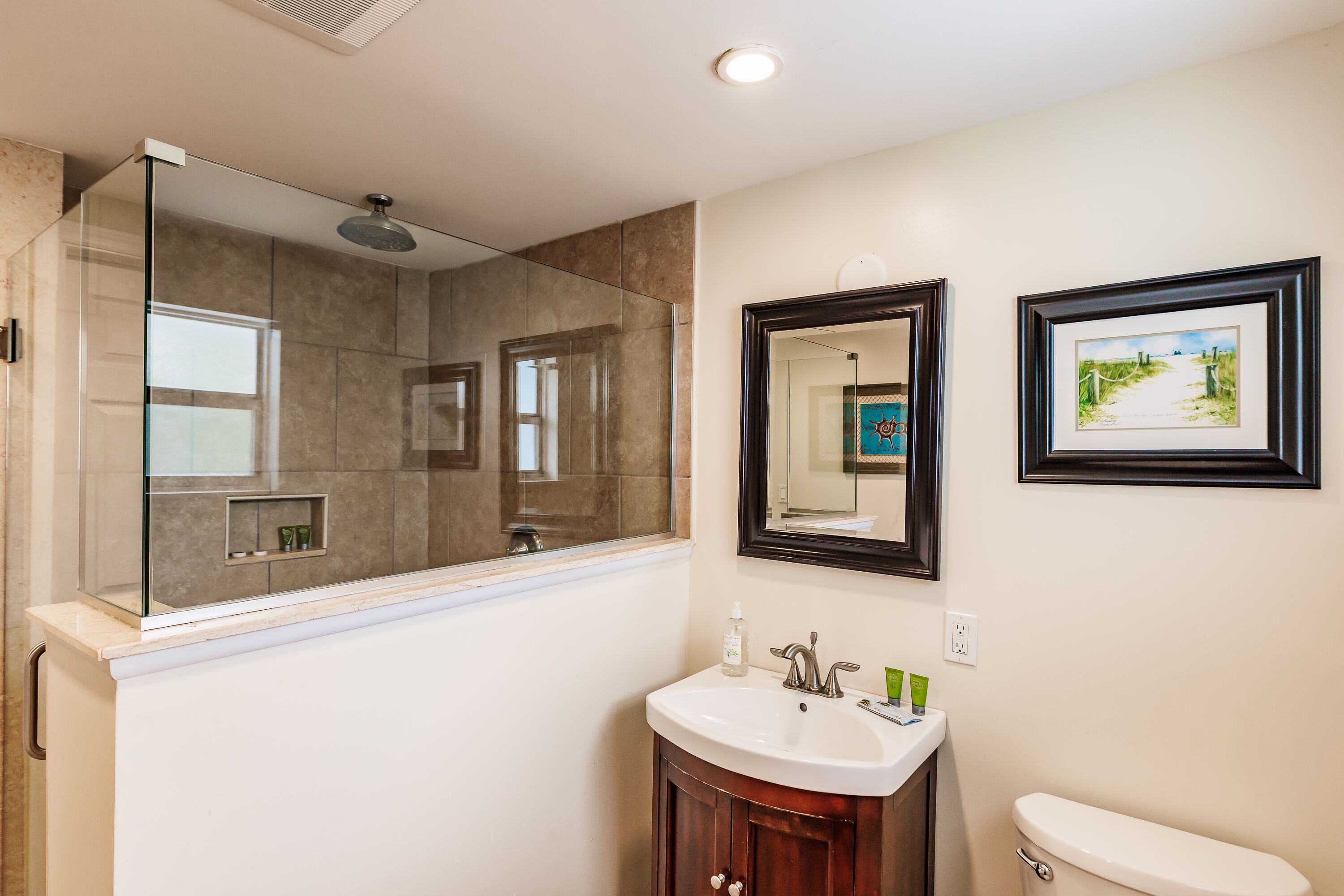 A bathroom with a glass shower enclosure, a white sink with a mirror above it, and a framed picture on the wall.