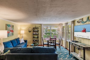 A living room with a blue sofa, a dining table with chairs, a bookshelf, and a TV displaying a beach scene.