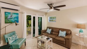 Ein Wohnzimmer mit braunem Ledersofa, einem türkisfarbenen Sessel, einem Glastisch, einem Deckenventilator und Blick ins Freie durch eine Schiebetür.