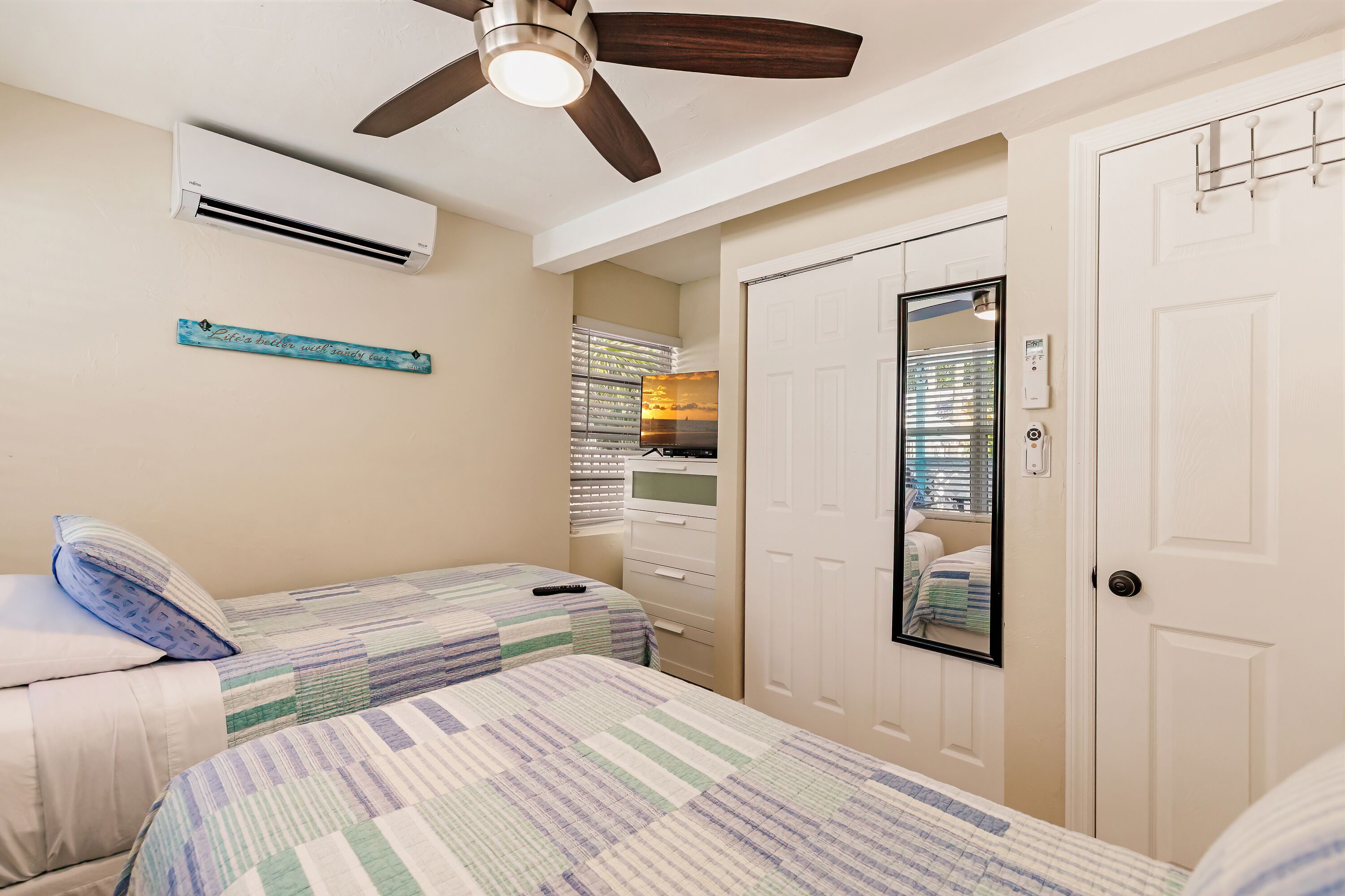 Quarto com duas camas, um ventilador de teto, um aparelho de ar condicionado, um espelho e uma porta branca.