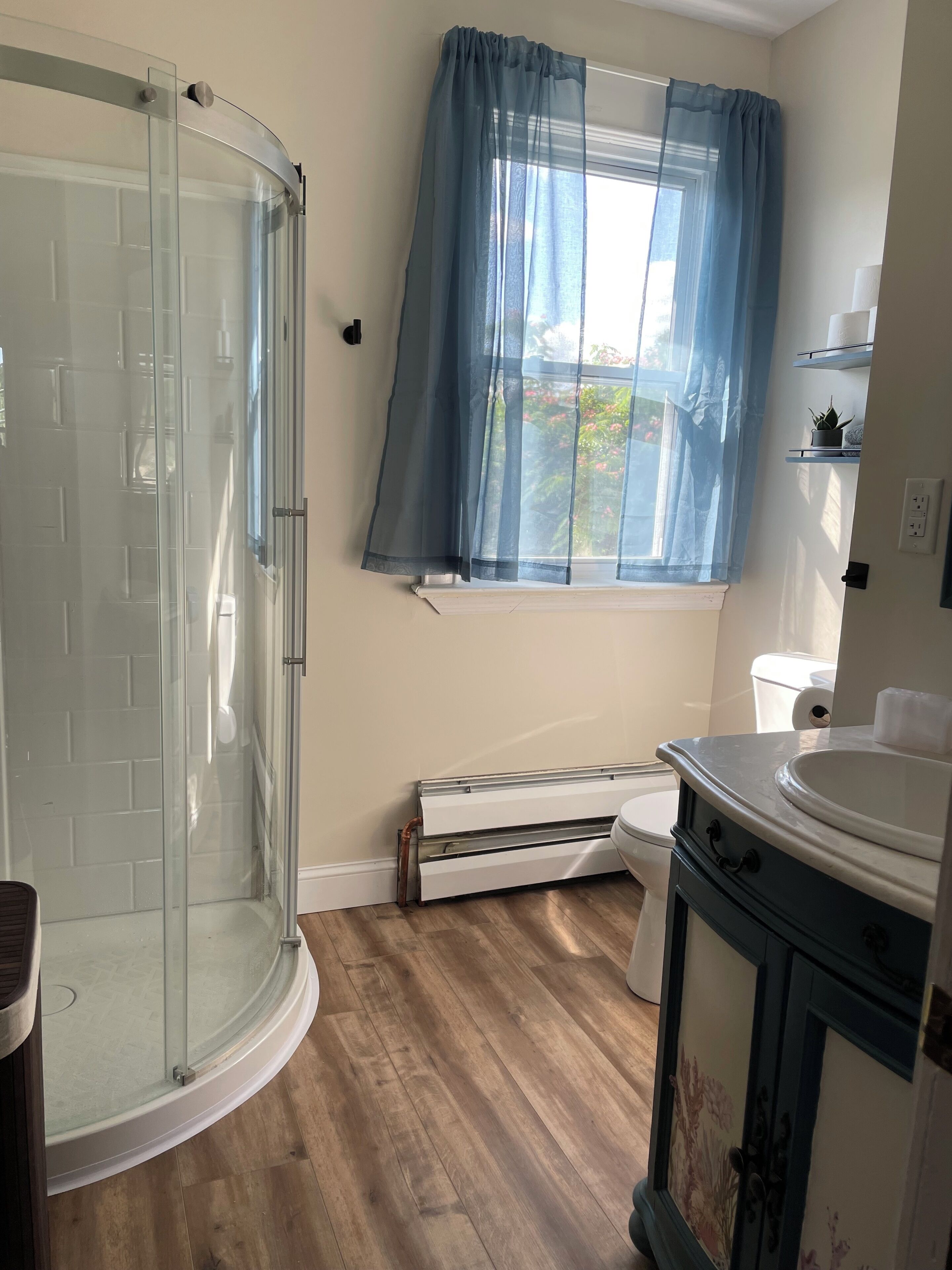 Un baño con cabina de ducha de vidrio, una ventana con cortinas azules, inodoro blanco y un lavabo con mueble decorativo.