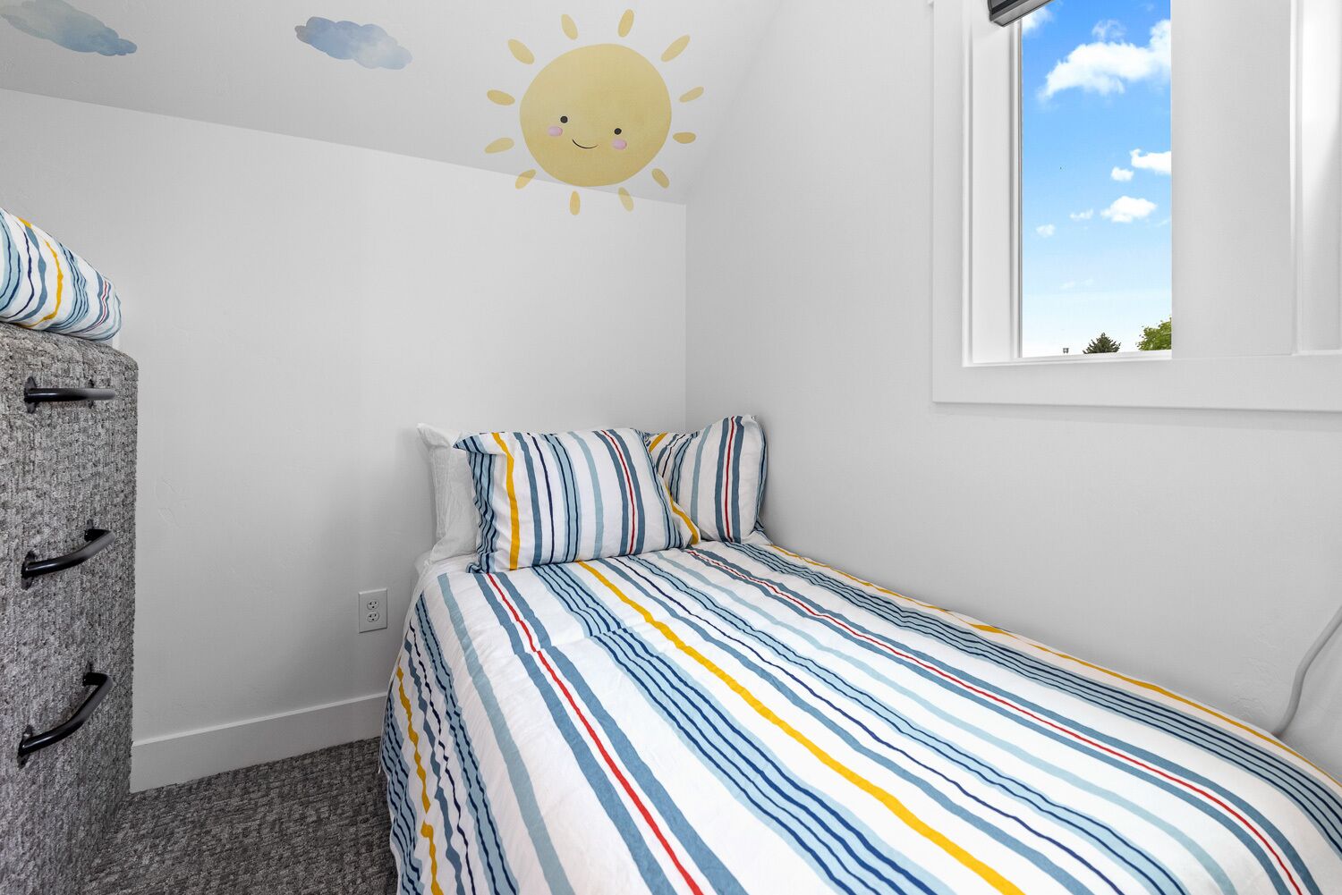 A bedroom with a striped bedspread, a dresser with black handles, and a wall decal of a smiling sun.