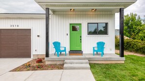 Una casa moderna de dos pisos con una puerta verde, dos sillas Adirondack azules y un garaje.