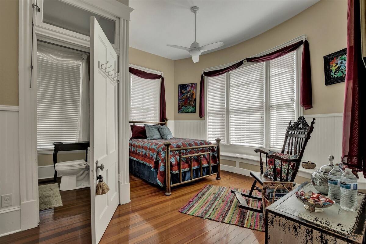 A bedroom with a bed, a chair, a table with items, and a window with curtains.