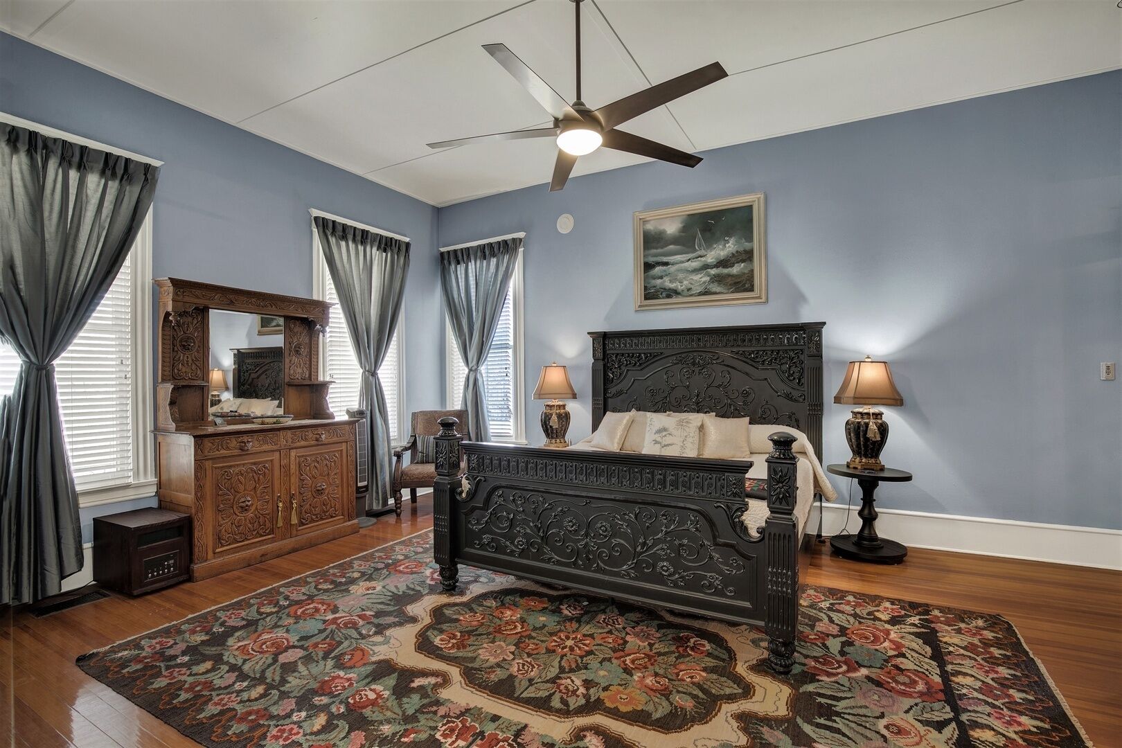 A bedroom with a large bed, a dresser, a ceiling fan, and a painting on the wall.