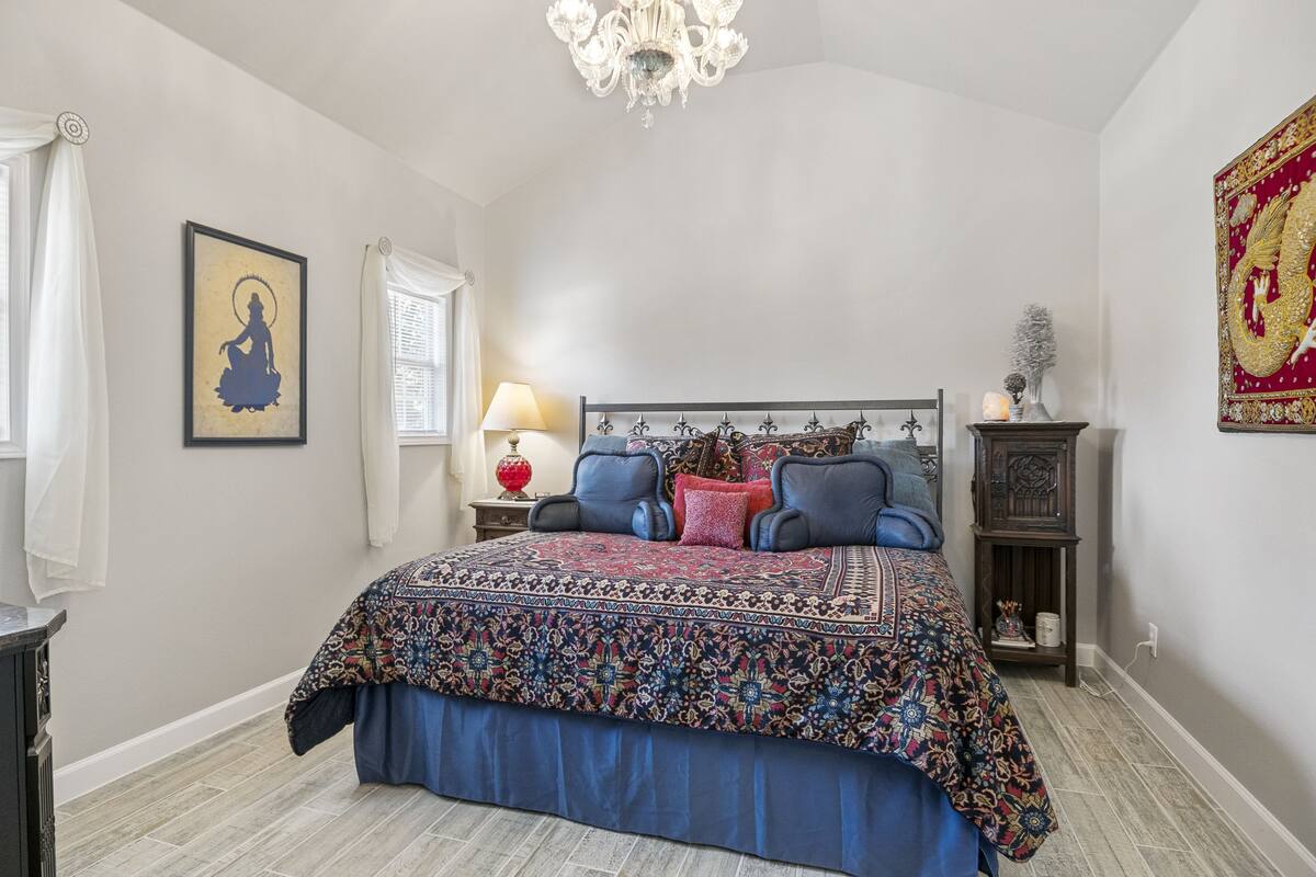 A bedroom with a large bed, a chandelier, a nightstand with a lamp, and a decorative wall piece.