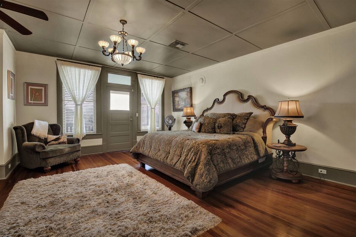 A bedroom with a large bed, a chair, a rug, a chandelier, and a door with curtains.