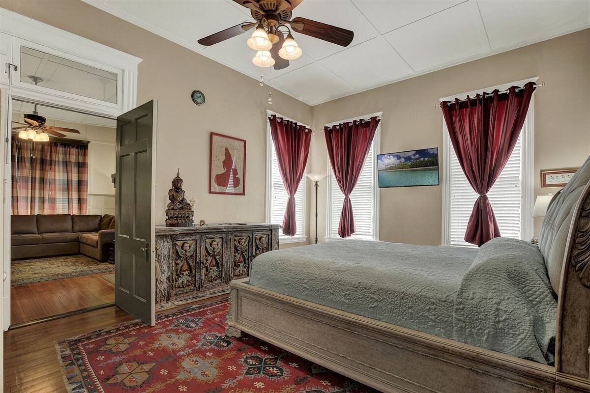 A bedroom with a bed, a dresser, a ceiling fan, and a view of the outdoors through the windows.