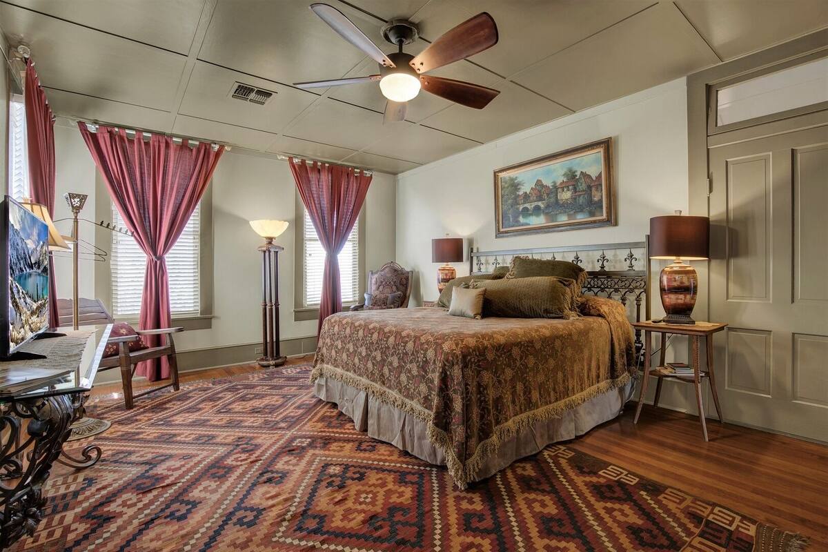A bedroom with a large bed, a ceiling fan, a painting on the wall, and a desk with a lamp.