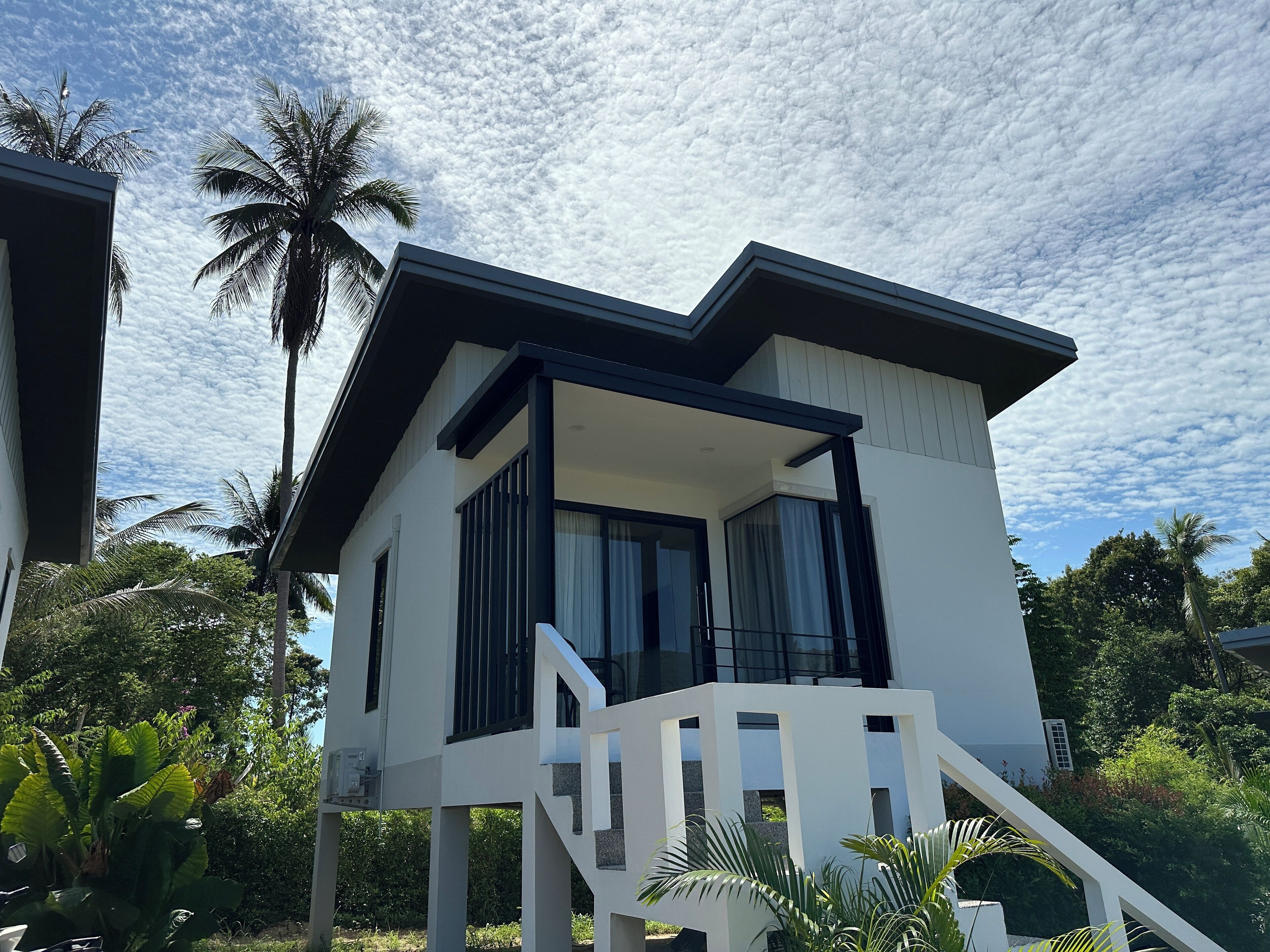 Uma casa moderna de dois andares com varanda, cercada por vegetação exuberante e palmeiras.
