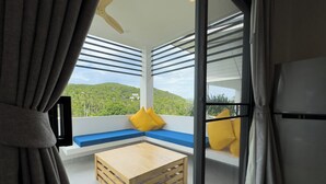 A balcony with a wooden bench and cushions, a small wooden table, and a view of a green hillside. - Naiwok Hills (Ko Pha-ngan)