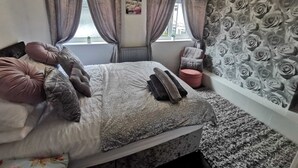 A bedroom with a bed, pillows, a window with curtains, a radiator, a wall with a floral pattern, and a ceiling light. - 2-Bed House in Macclesfield Cheshire (Macclesfield)