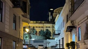 A narrow street at night with buildings on either side, a parked car, and a building with a clock tower in the background. - Opera Apartment Sleeps 4 with Patio (T'bilisi)