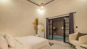 A modern bedroom with a bed, a sofa, a small table, and a sliding glass door leading to a balcony with a pool.