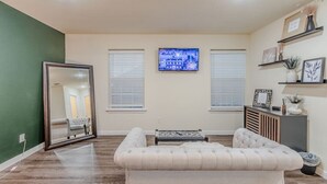 Una sala de estar con un espejo grande, dos ventanas con persianas, un televisor y un sofá.