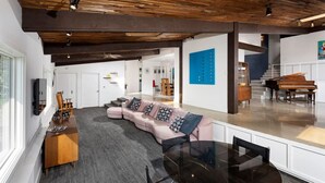 A spacious living area with a curved pink sofa, a glass coffee table, and a wooden side table. A staircase leads to a piano area.