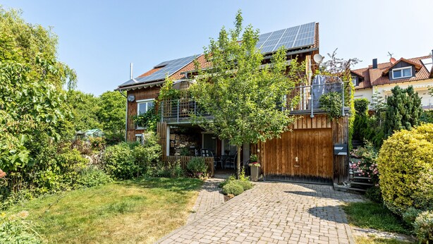 Et moderne hus med solpaneler, træfacade og en belagt indkørsel, der fører til et overdækket udendørs område.