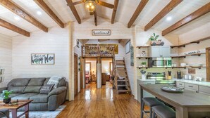 Interior acogedor de una cabaña con techo de madera, un sofá con almohada que dice 'Stay', una zona de cocina y una escalera que lleva a un dormitorio en la buhardilla.