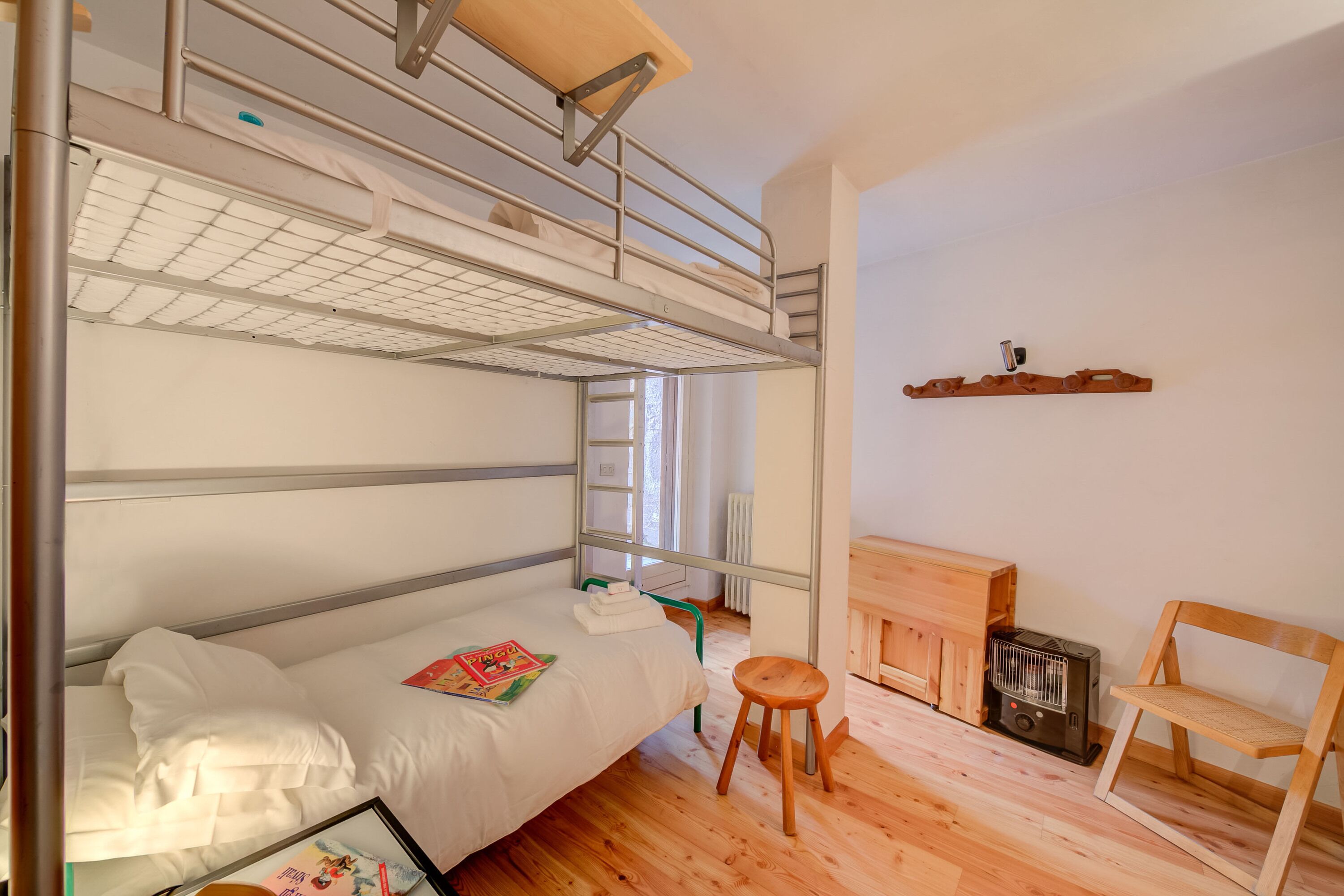 A bunk bed room with a wooden floor, a small stool, a chair, a radiator, and a shelf with a heater.