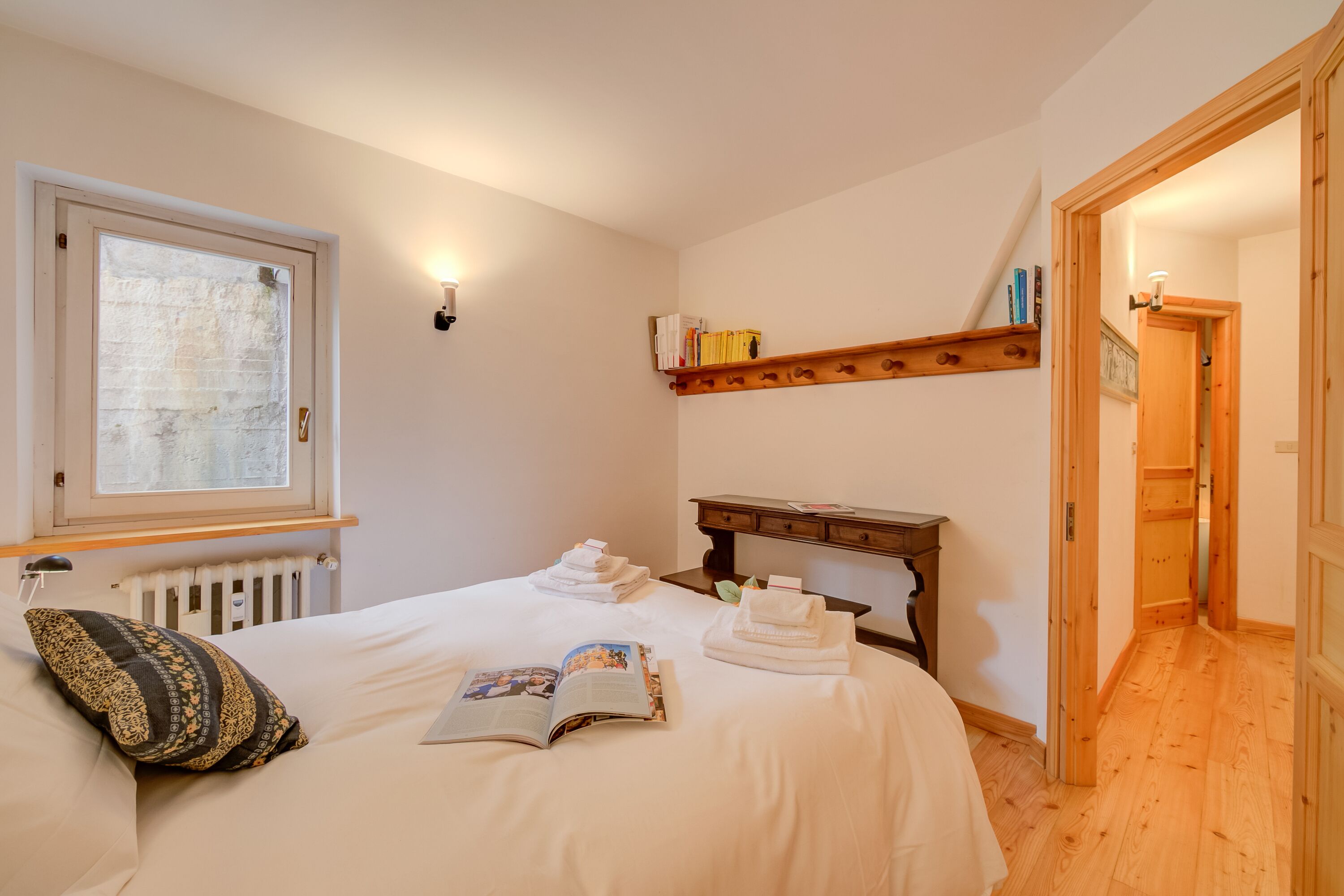 Cama bem arrumada com um livro, toalhas e um travesseiro. Há uma estante de madeira com livros e um radiador acima da cama.