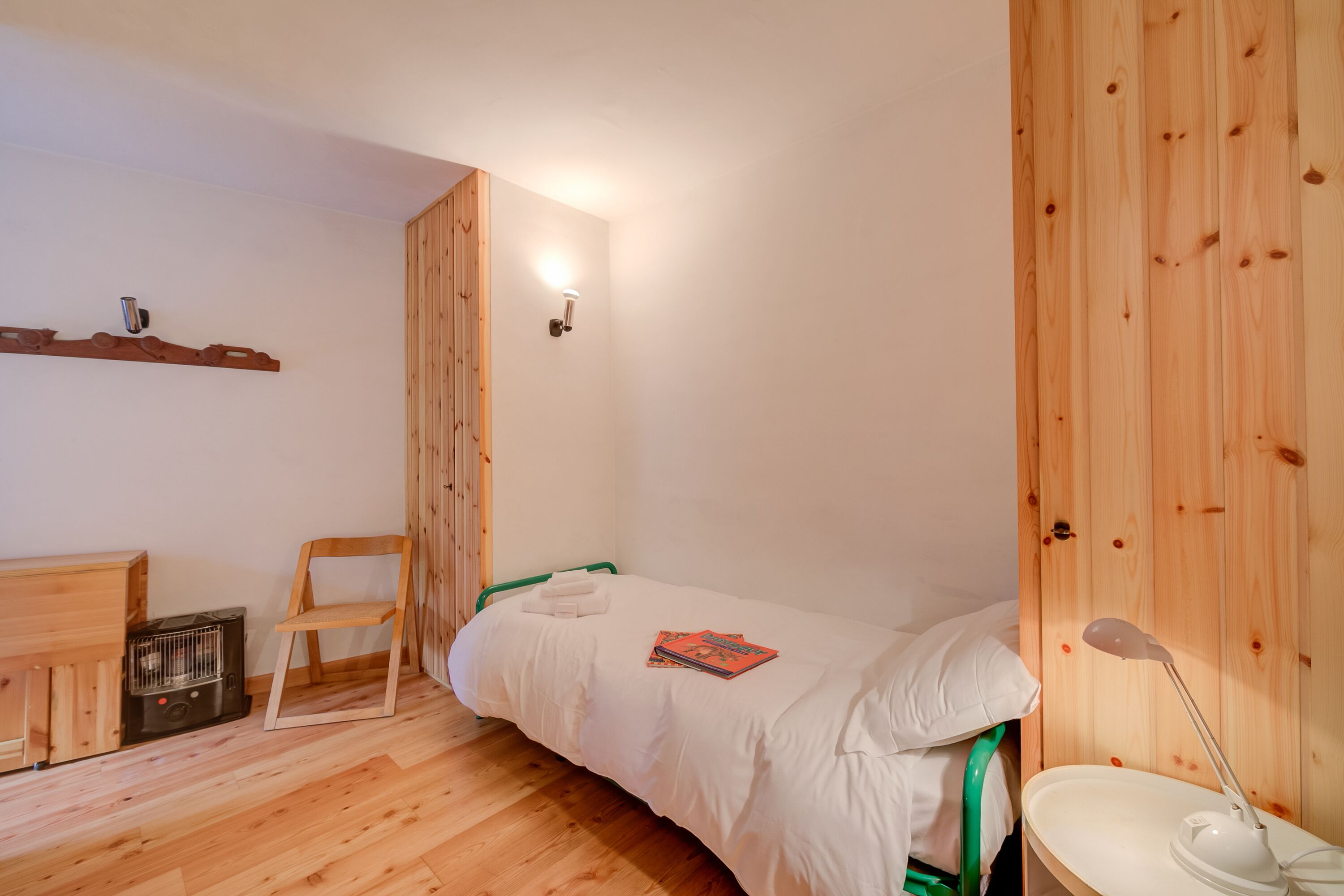 Een kleine, knusse kamer met een houten vloer, een enkel bed met wit beddengoed, een groene stoel, een klein bureau met een verwarming en een wandlamp.