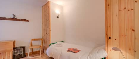 Een kleine, knusse kamer met een houten vloer, een enkel bed met wit beddengoed, een groene stoel, een klein bureau met een verwarming en een wandlamp.