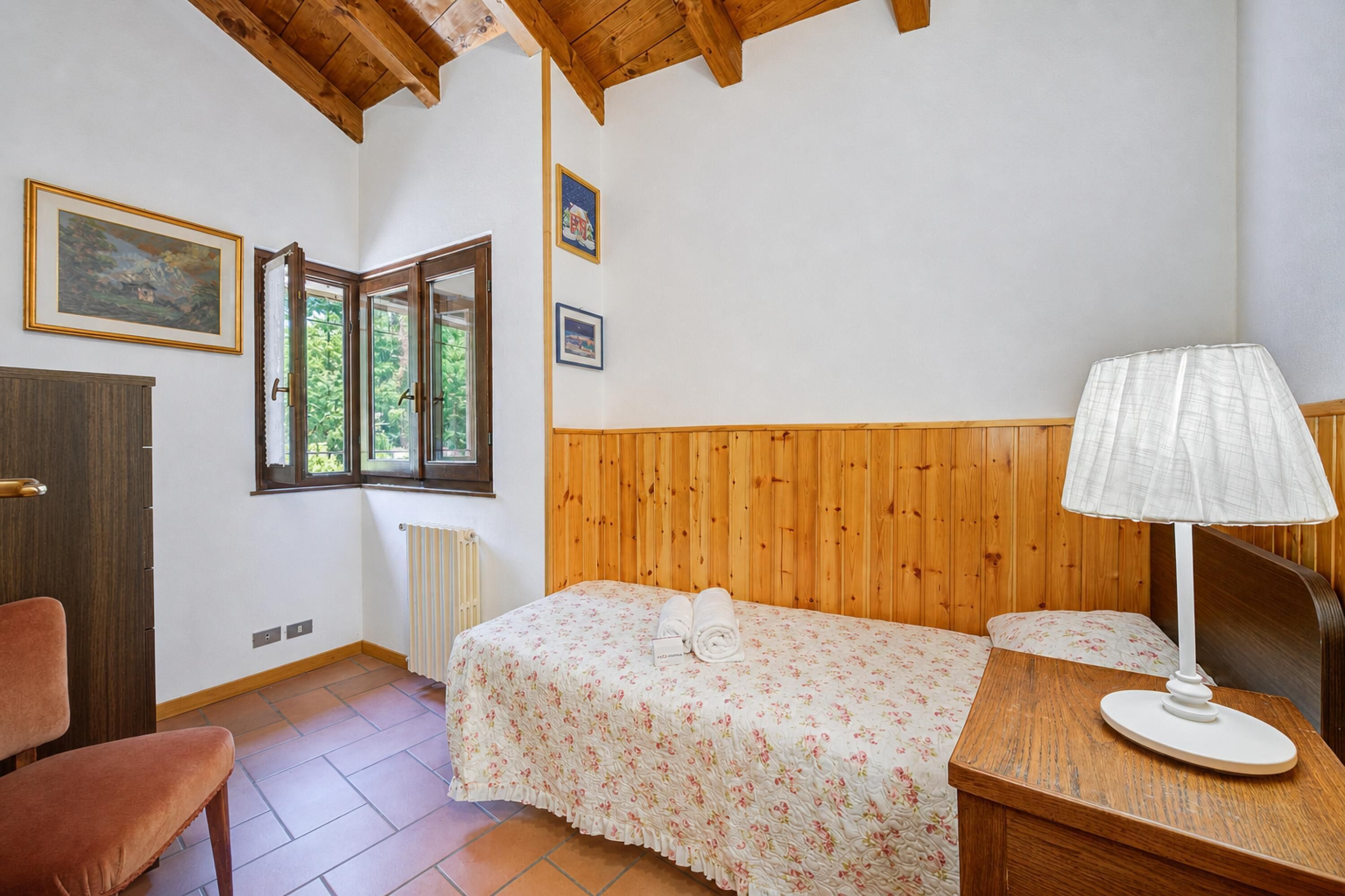 Ein Schlafzimmer mit einem hölzernen Kopfteil, einem Bett mit floralem Überwurf, einem Nachttisch aus Holz mit einer Lampe, einem Fenster mit Blick ins Grüne und einem gerahmten Bild an der Wand.