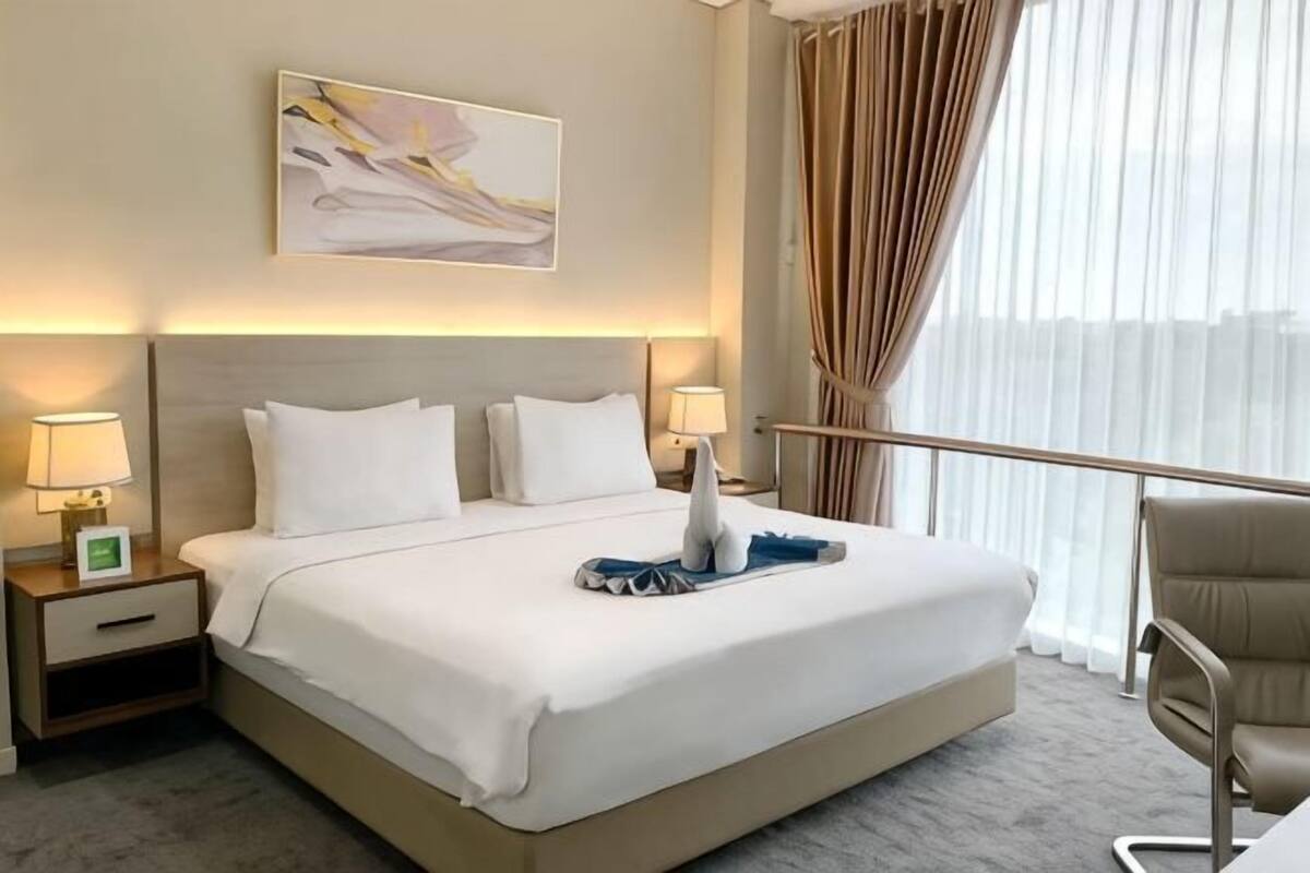 A hotel room with a large bed, two bedside tables with lamps, a chair, a window with curtains, and a painting on the wall.