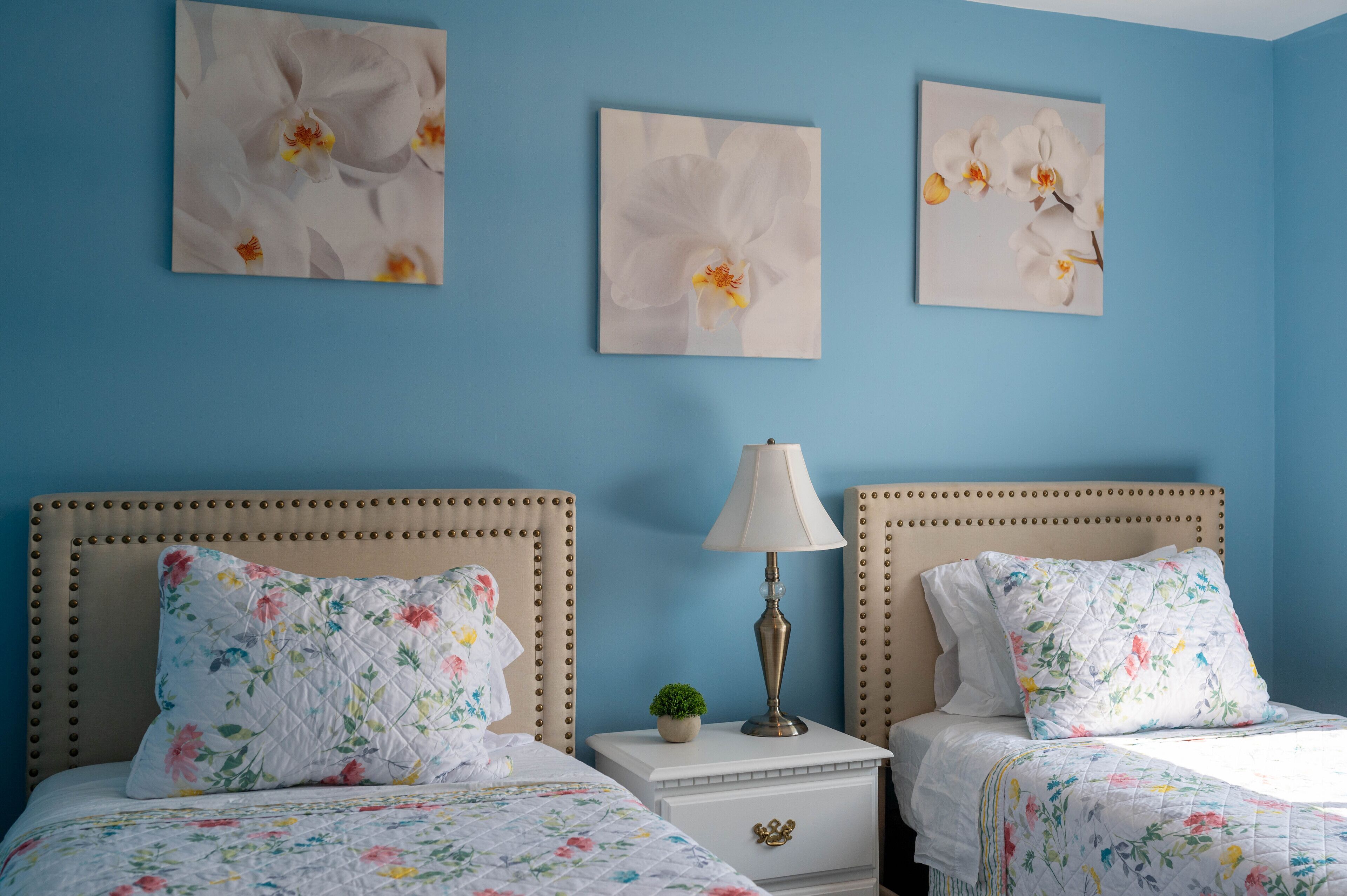 A bedroom with two beds, floral bedding, a bedside table with a lamp, and three orchid paintings on the wall.