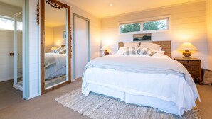 A bedroom with a large bed, two bedside tables with lamps, a large mirror, and a window with a view of trees. - Oriwa Villa Oneroa near Mudbrick (Waiheke Island)