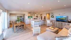 A spacious living area with a dining table, white wicker chairs, a sofa, and a TV. - Oriwa Villa Oneroa near Mudbrick (Waiheke Island)