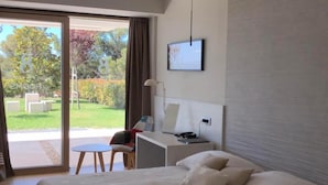 Ein modernes Hotelzimmer mit einem großen Bett, einem Schreibtisch, einem Sessel, einem Fernseher und einer Glastür, die auf eine Terrasse führt.