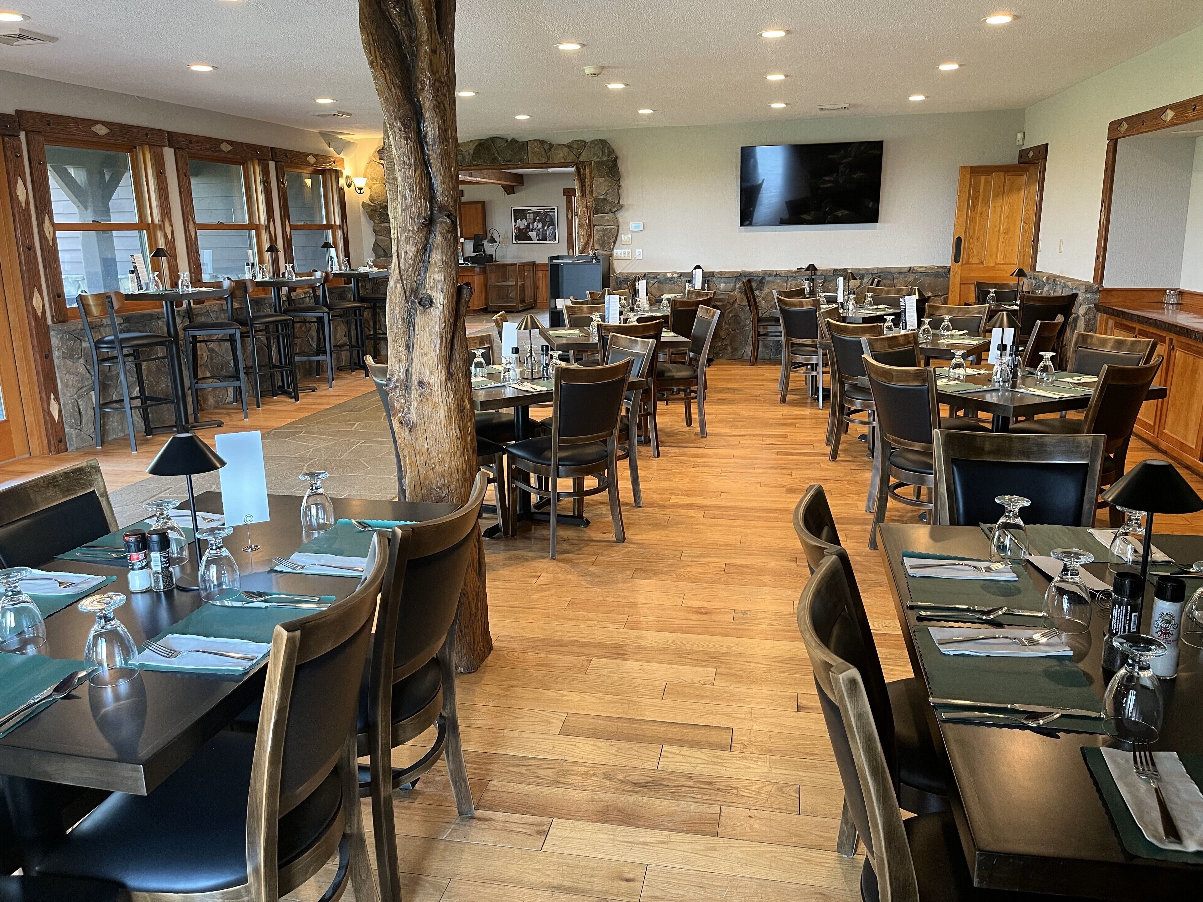 A restaurant with multiple dining tables and chairs, wooden floors, stone walls, and a large tree in the center.