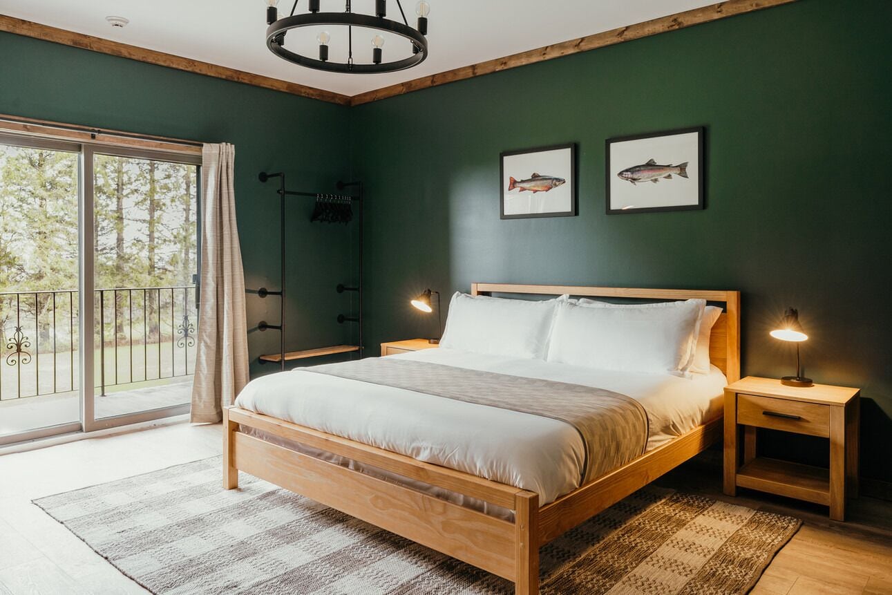 Une chambre à coucher avec un lit en bois, deux tables de chevet avec des lampes, une grande fenêtre donnant sur des arbres et deux tableaux représentant des poissons, encadrés et accrochés au mur.