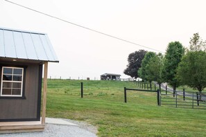 A small, single-story building with a metal roof, a fenced area, and a grassy hill in the background.