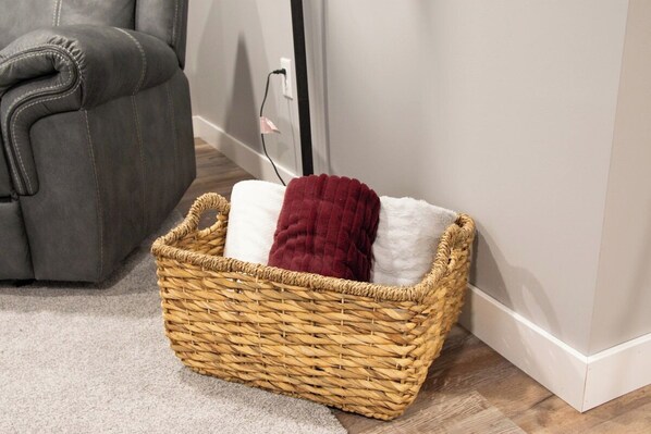 A wicker basket with a white and a maroon cushion, placed on a carpeted floor next to a gray sofa.