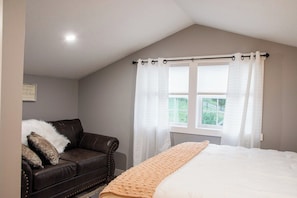 A bedroom with a bed, a window with curtains, a brown leather sofa, and a wall-mounted picture.