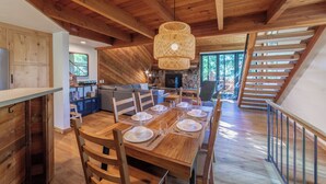 Une salle à manger spacieuse avec une longue table en bois, dressée pour quatre personnes, une cheminée en pierre et un escalier menant à un niveau supérieur.