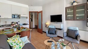 Een moderne woonkamer met een bank, fauteuils, een salontafel en een televisie. In de achtergrond is een keuken zichtbaar met witte kastjes en een magnetron.