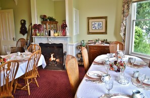 A dining room with a fireplace, wooden chairs, a table set for a meal, and a window with a view of greenery.